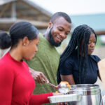 Our Projects A closeup shot of a man pouring food into a plate standing between two women