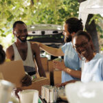 Our Projects African American woman and caucasian man smile while providing boxes of canned goods handing out donation boxes to needy. Individuals volunteering by sharing necessities to poor and homeless people.