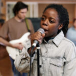 Our Projects Waist up portrait of young black woman singing to microphone while recording music with band in professional studio